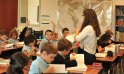 A teacher instructs in the middle of a class with a large atlas displaying all the places around the globe